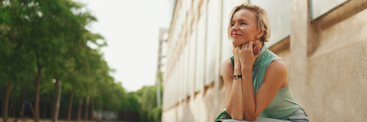 A woman with blonde hair sitting outdoors next to a beige building, wearing a sleeveless sage green top and light blue jeans, looking thoughtfully to the side.